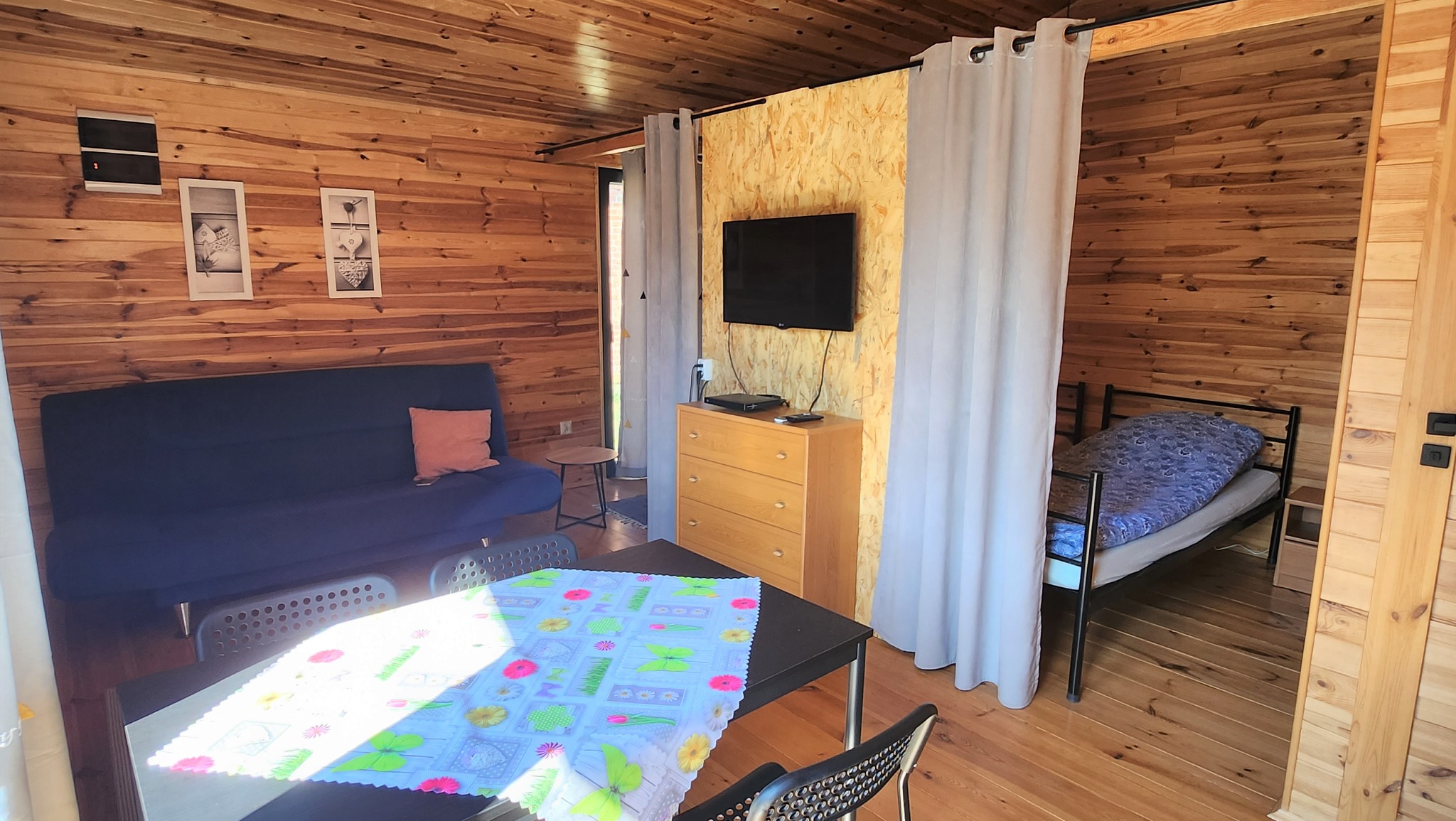 Drewniany domek letniskowy: salon z rozkładaną sofą, stolik z kolorowym obrusem, sypialnia oddzielona zasłoną, telewizor na komodzie.