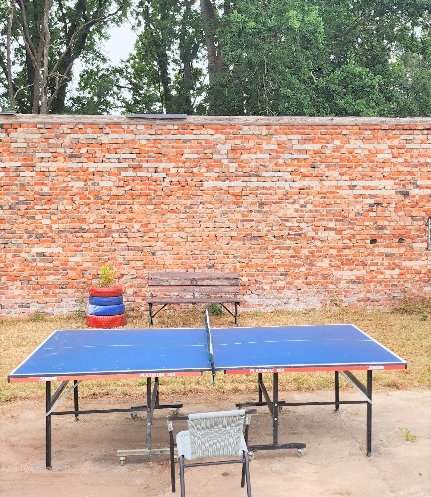 Stół do ping-ponga Platinum 200 ustawiony na zewnątrz, z krzesłem, ławką i dekoracją z opon na tle ceglanego muru.