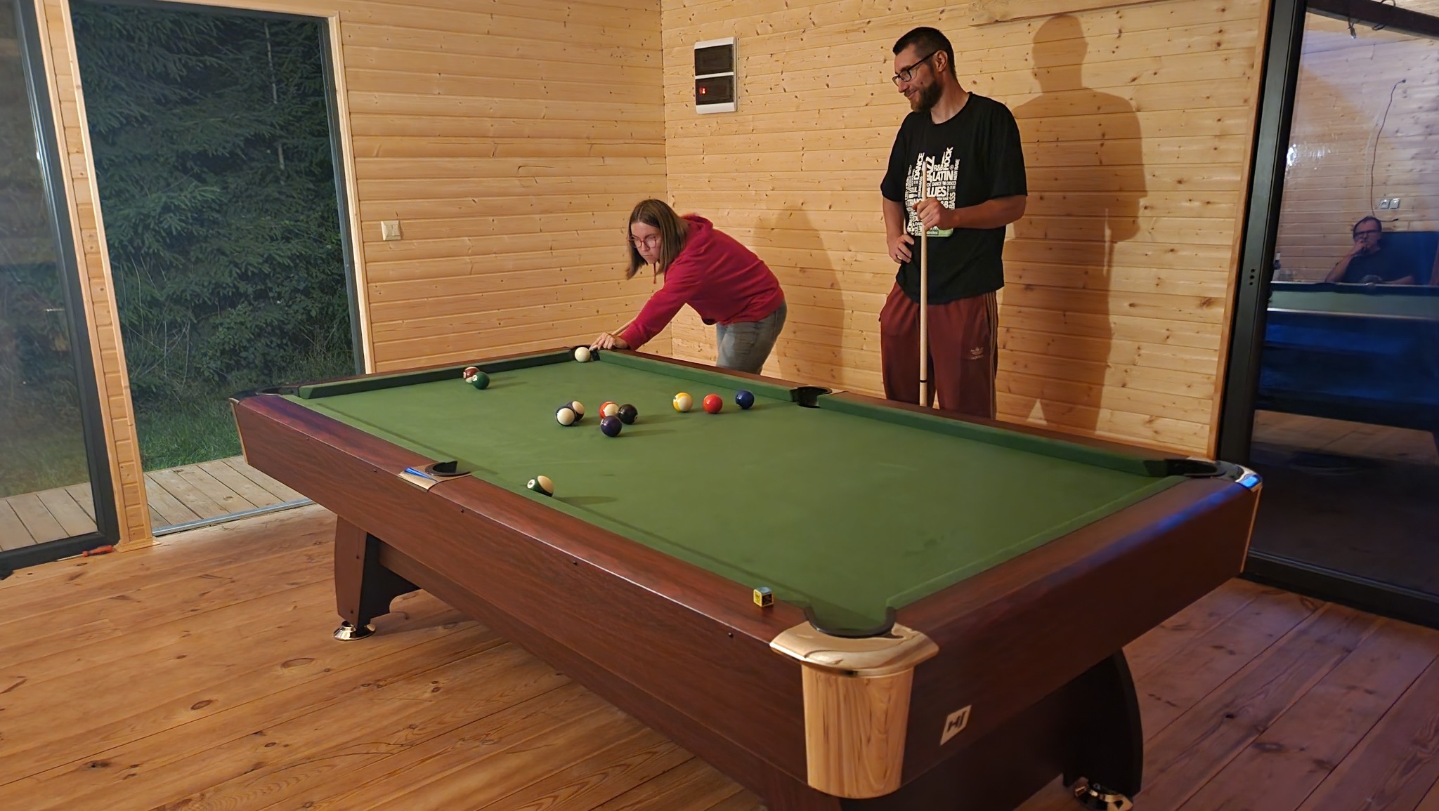 Wnętrze domku z drewnianą boazerią: kobieta celuje bilę na stole bilardowym, obok stoi mężczyzna z kijem, w tle widoczny basen za szklaną ścianą.