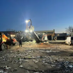 Praca maszyny sortującej odpady z podajnikiem taśmowym i dźwigiem w szaro-pomarańczowym oświetleniu zmierzchu, na placu z rozrzuconymi odpadami, w tle budynki.