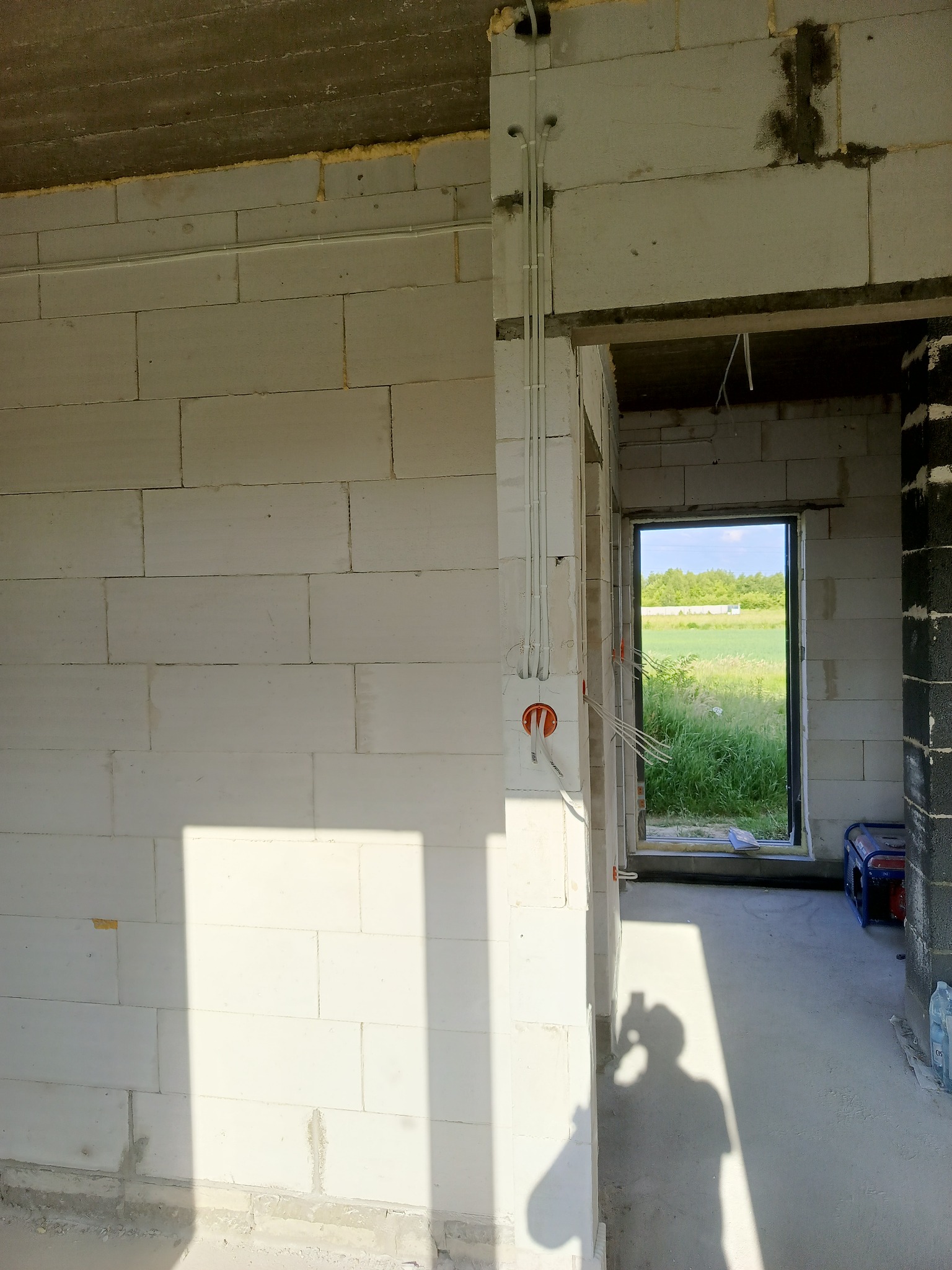 Wnętrze surowego budynku w trakcie prac instalacyjnych: widoczne przewody elektryczne prowadzone wzdłuż ściany z bloczków, puszka instalacyjna z pomarańczowymi elementami, cień osoby fotografującej...