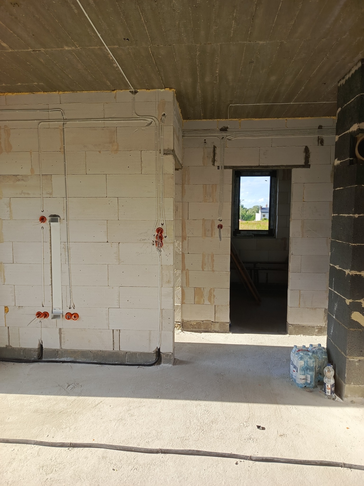 Surowy stan wnętrza budynku w trakcie instalacji elektrycznej: widoczne przewody w peszlach, puszki instalacyjne w ścianach z betonu komórkowego, betonowy strop, okno z widokiem na zewnątrz.