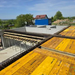 Wysoki kąt widzenia na plac budowy z widocznymi fundamentami, szalunkiem z żółtych desek Doka 3-SO i wystającą stalą zbrojeniową, z domami w tle pod błękitnym niebem.