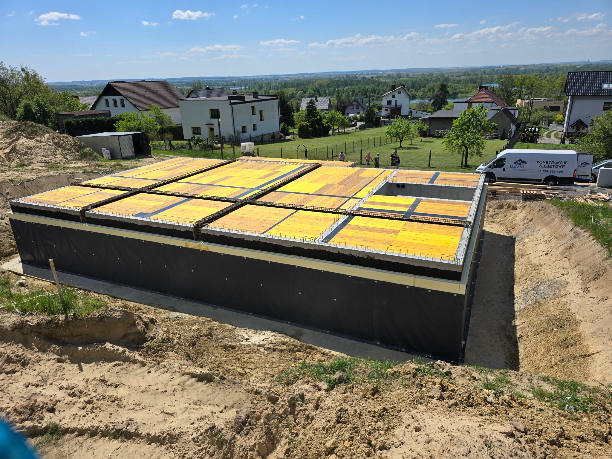 Zabezpieczony wykop pod budynek z deskowaniem i żółtymi płytami szalunkowymi na górze, widoczny samochód dostawczy firmy budowlanej w tle.