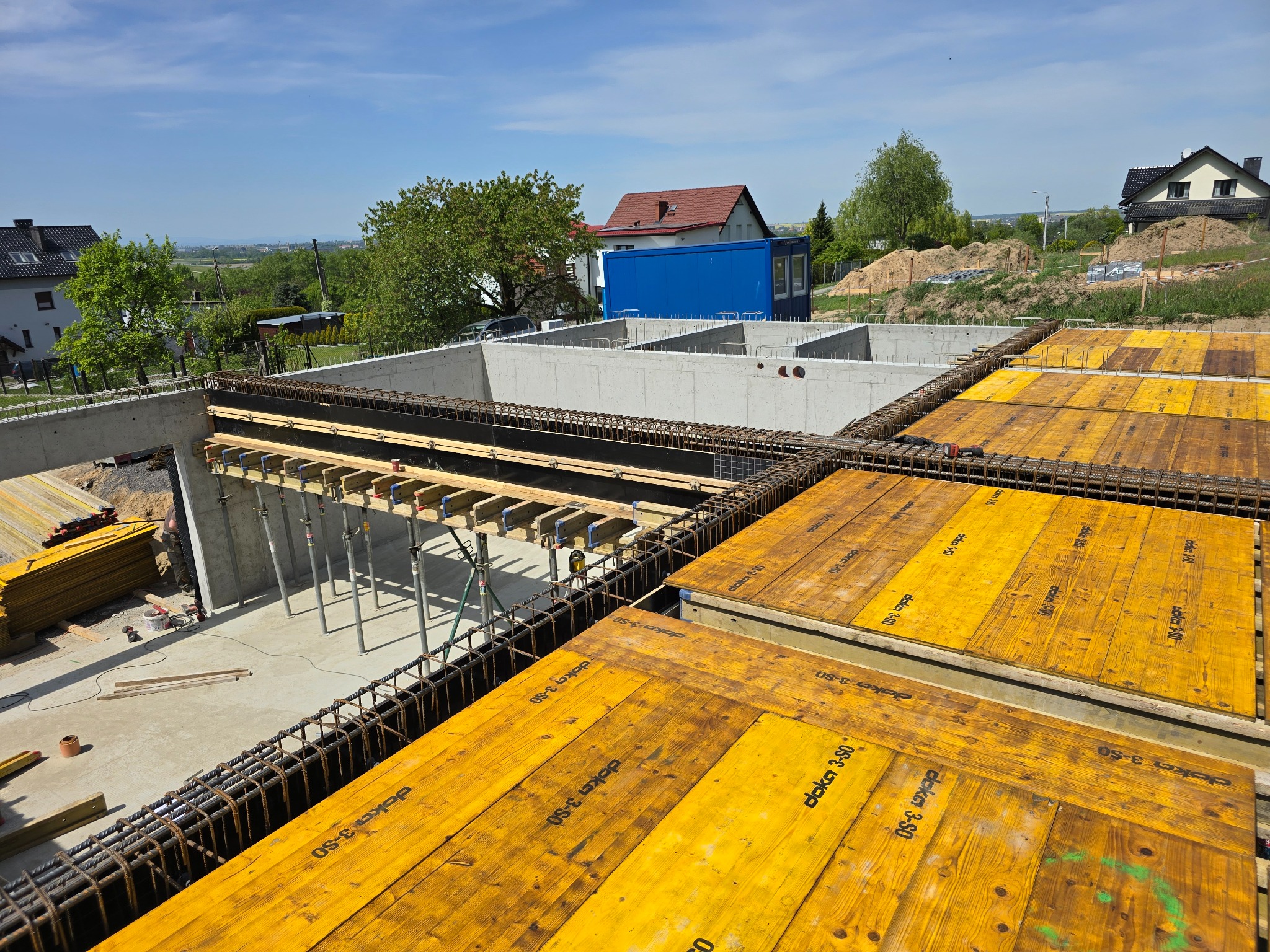 Wysoki kąt widzenia na plac budowy z widocznymi fundamentami, szalunkiem z żółtych desek Doka 3-SO i wystającą stalą zbrojeniową, z domami w tle pod błękitnym niebem.