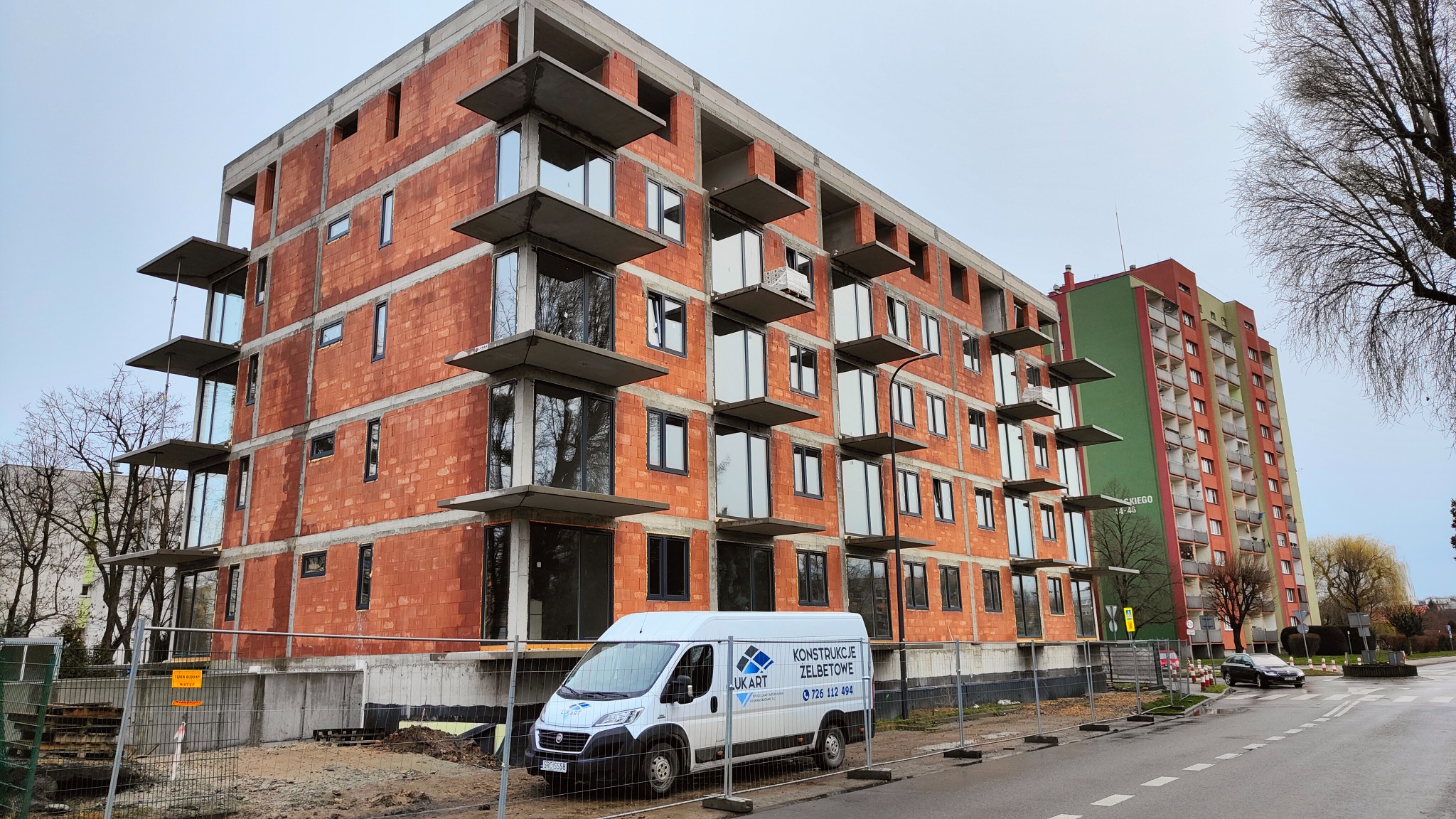 Wielopiętrowy budynek w trakcie budowy z czerwonej cegły, widoczne betonowe elementy balkonów, okna wstawione, na pierwszym planie samochód dostawczy z logo firmy budowlanej.