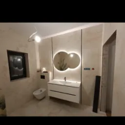 Nowoczesna łazienka z beżowymi ścianami, białym wiszącym bidetem i szafką pod umywalkę z dwiema szufladami, nad którą wiszą dwa okrągłe lustra z podświetleniem LED.