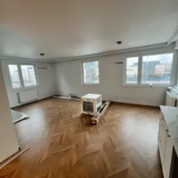 Salon po remoncie z parkietem ułożonym w jodełkę, białe ściany, okna z widokiem na miasto, kartony z panelami pod ścianą i nowoczesne lampy sufitowe.