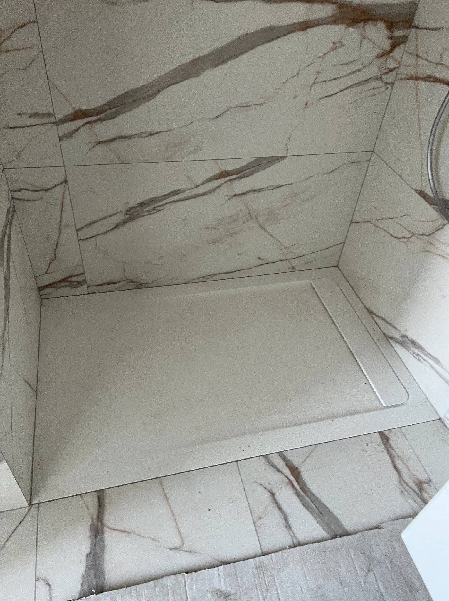 Płytki imitujące marmur na ścianach i podłodze kabiny prysznicowej z białym brodzikiem. Widoczne fugi i fragment odpływu.