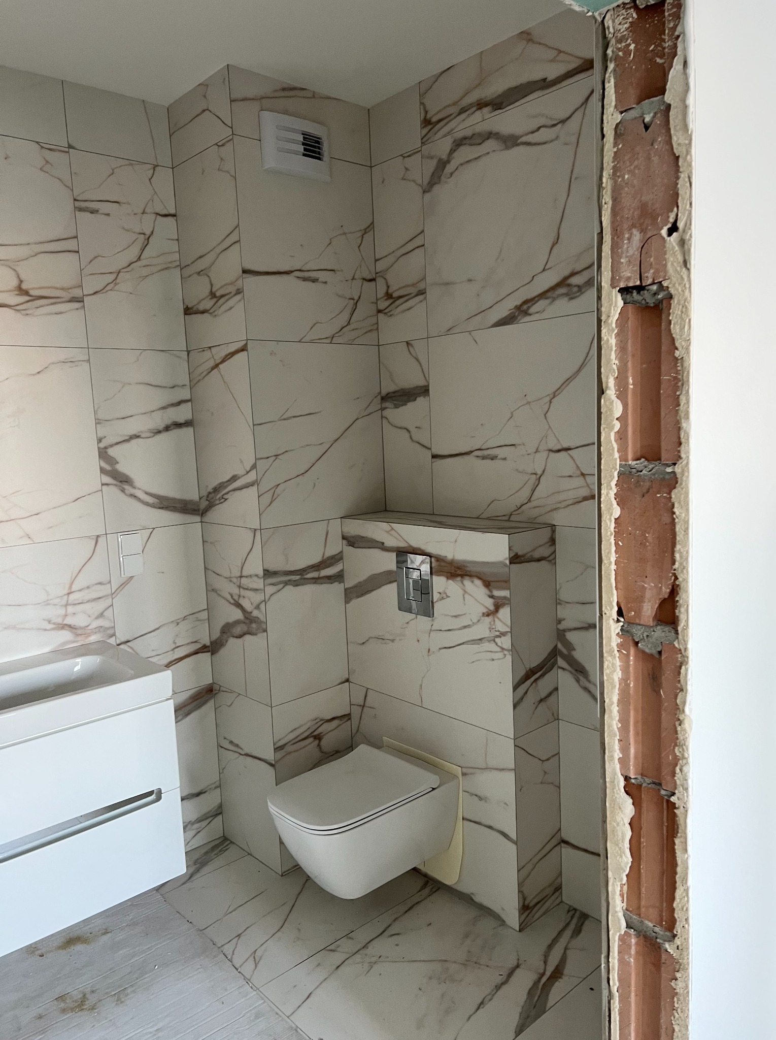 Wykończona łazienka z białymi płytkami imitującymi marmur, wisząca toaleta i umywalka z szafką, widoczny fragment surowej ściany z cegły.