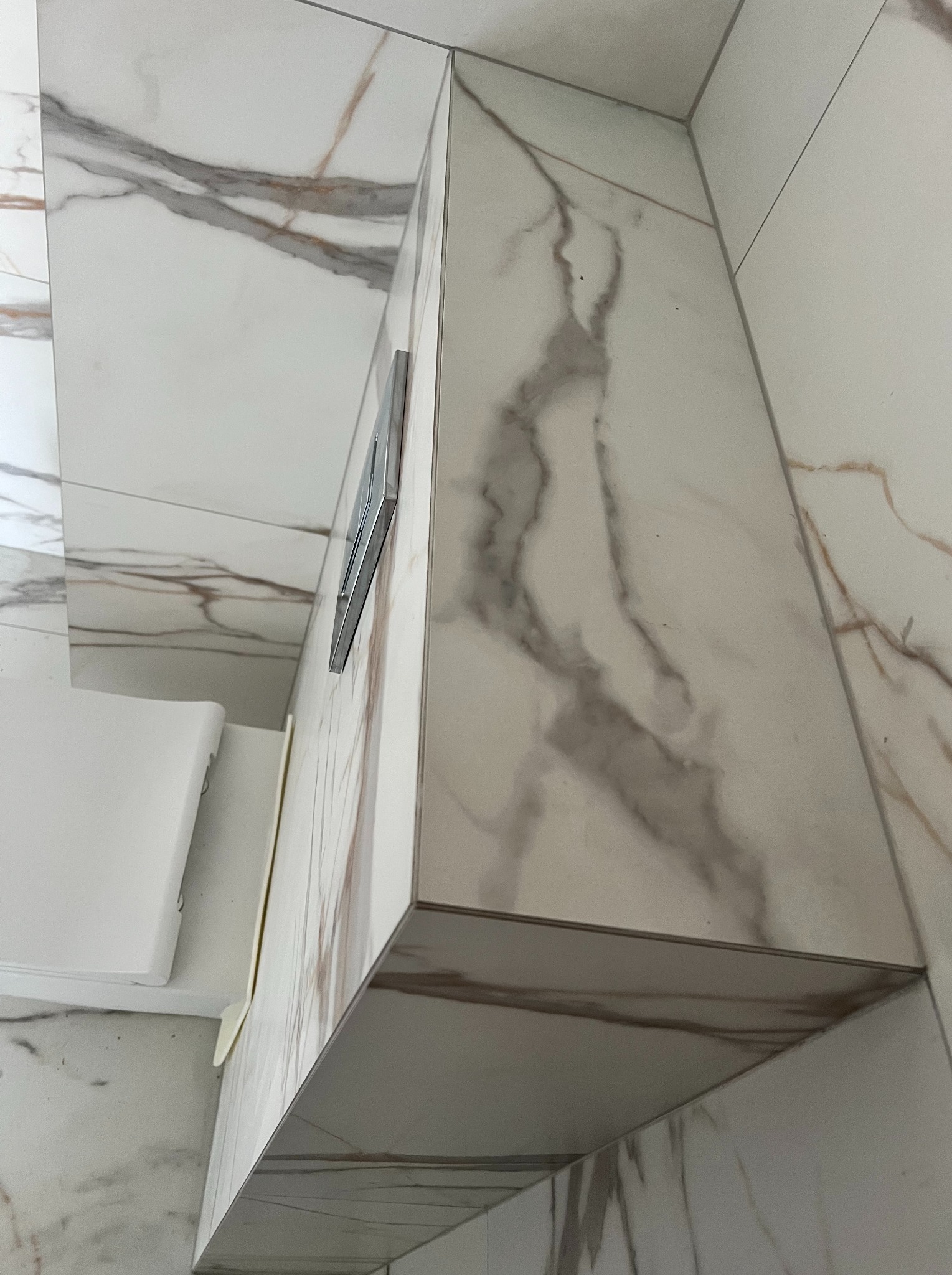 Detale wykończenia toalety: białe płytki imitujące marmur z szarymi żyłami, obudowa stelaża podtynkowego, chromowany przycisk spłukujący.