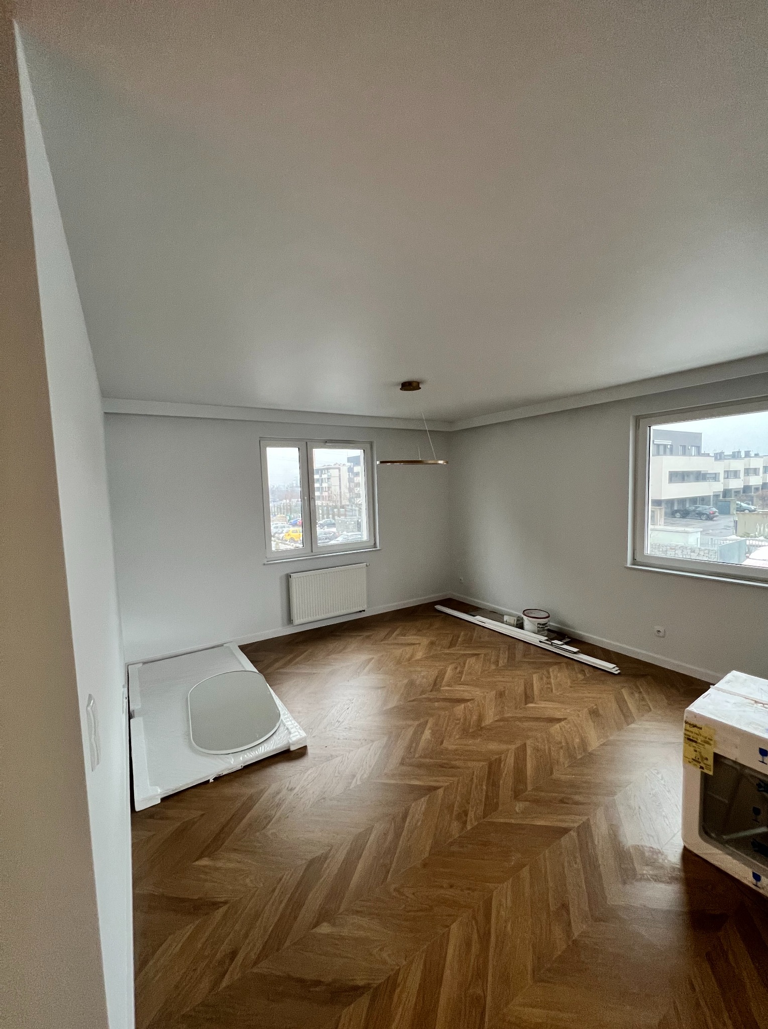 Pusty pokój z parkietem ułożonym we wzór jodełki, świeżo pomalowanymi białymi ścianami, dwoma oknami z widokiem na miasto i elementami przygotowanymi do dalszych prac wykończeniowych.