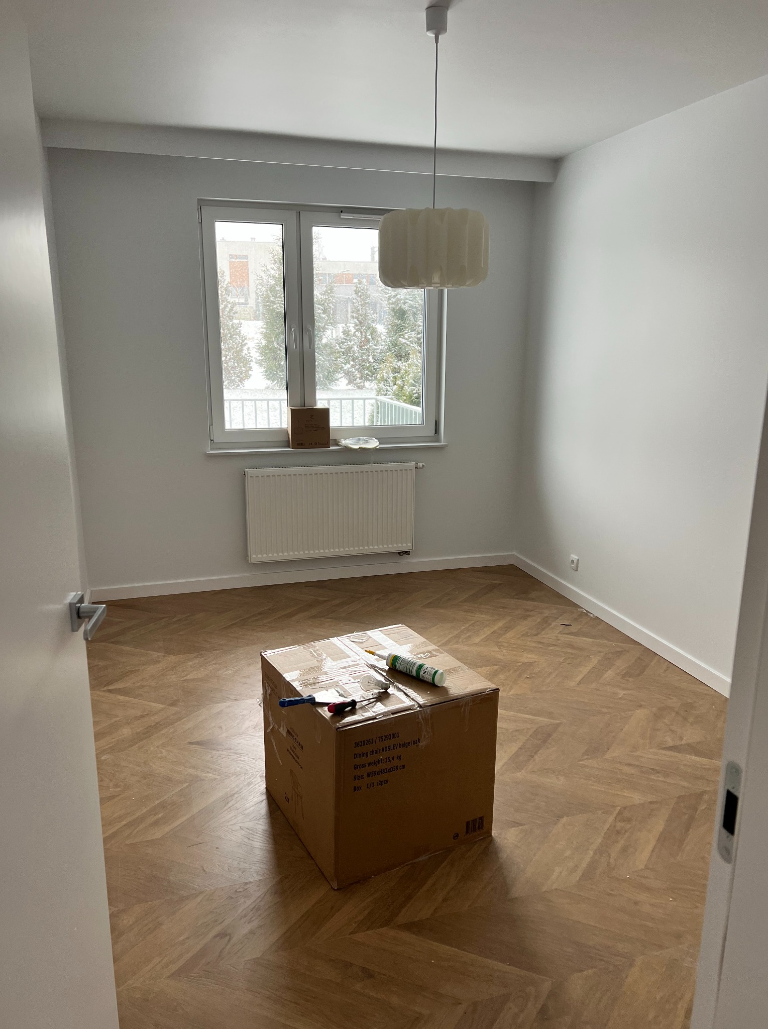 Pokój po remoncie z nową podłogą w jodełkę, karton z narzędziami na środku, białe ściany i okno z widokiem na zimowy krajobraz.