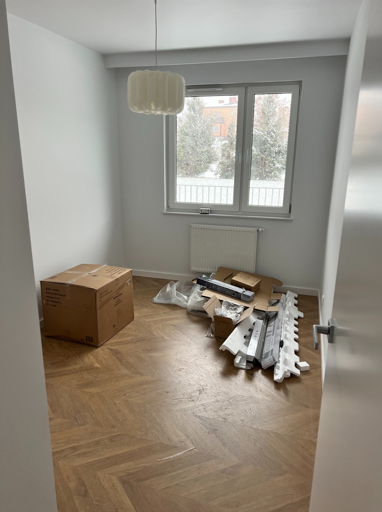 Pusta, jasna przestrzeń z parkietem w jodełkę, kartonowym pudłem i rozpakowanymi elementami mebli pod oknem, widok na zimowy krajobraz.