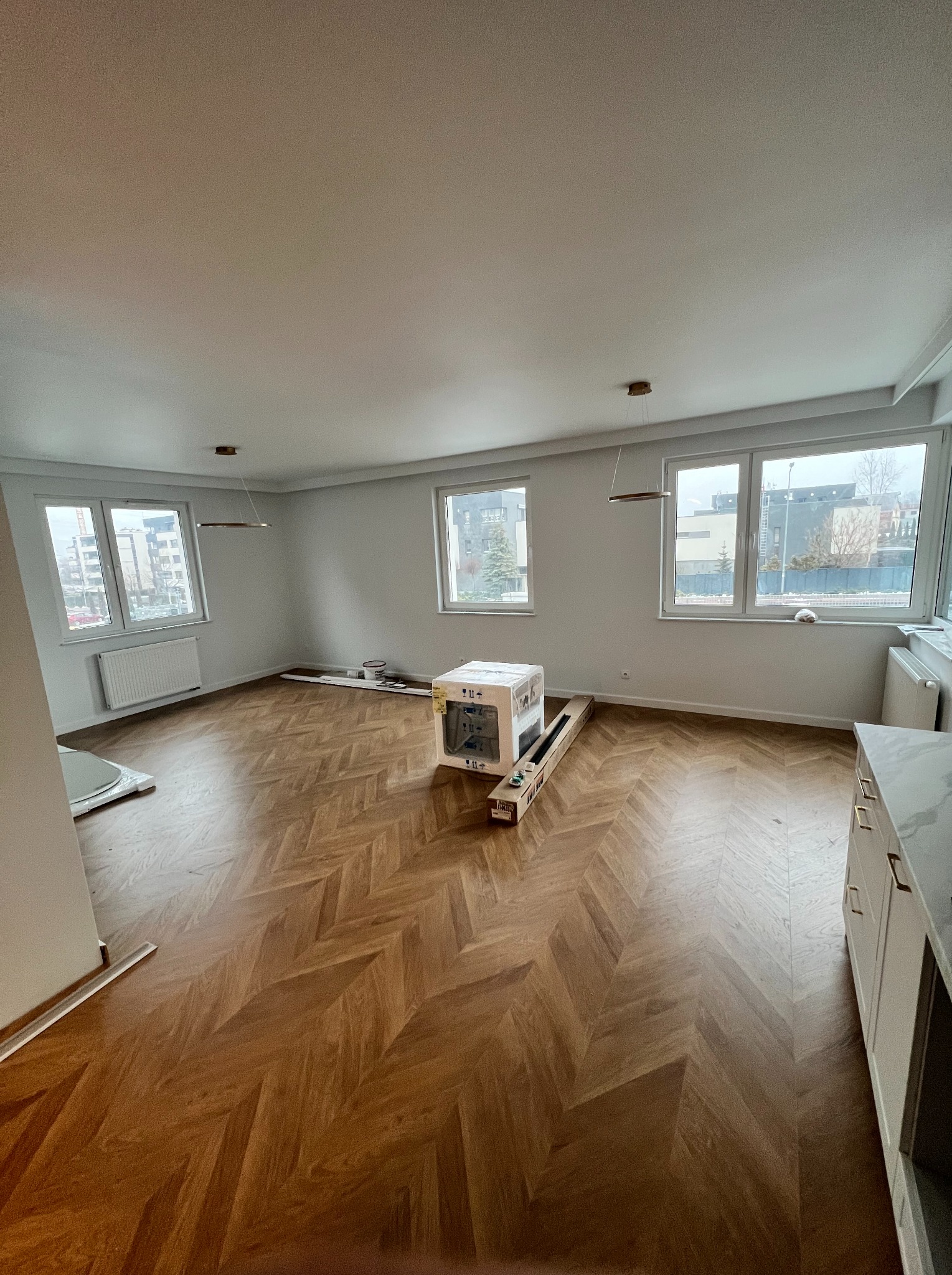 Salon po remoncie z parkietem ułożonym w jodełkę, białe ściany, okna z widokiem na miasto, kartony z panelami pod ścianą i nowoczesne lampy sufitowe.