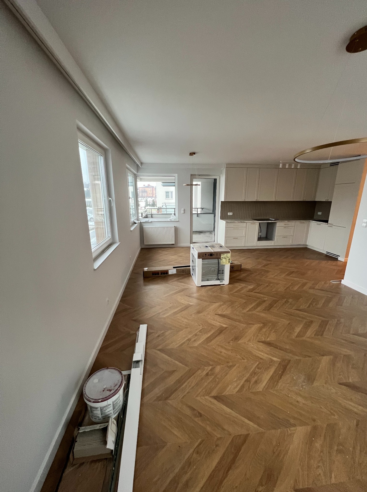 Nowoczesne wnętrze mieszkania w trakcie prac wykończeniowych; widoczna drewniana podłoga ułożona w jodełkę, białe ściany, okna, zabudowa kuchenna w tle oraz materiały budowlane i puszka farby przy...