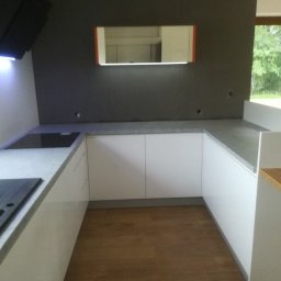 House Of Emma - Nowoczesna kuchnia w kształcie litery U z białymi, lakierowanymi frontami, szarym blatem i drewnianą podłogą. Nad płytą grzewczą zamontowany czarny okap z niebieskim podświetleniem LED. Na ścianie...