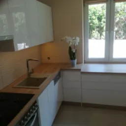 Nowoczesna kuchnia z białymi, lakierowanymi frontami, drewnianym blatem i płytkami z falistym wzorem na ścianie, z oknem i orchideą.