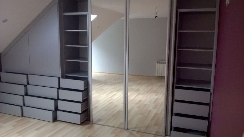Szara szafa wnękowa z lustrzanymi drzwiami przesuwnymi, dopasowana do skosu dachu, z licznymi szufladami i półkami, widok na puste pomieszczenie z parkietem i grzejnikiem.