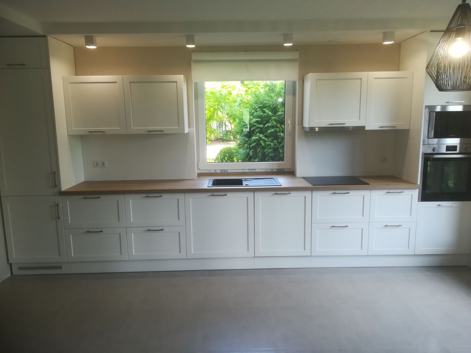 Jasna kuchnia z białymi szafkami na wymiar, drewnianym blatem i widokiem na zieleń przez okno, oświetlona punktowymi lampami sufitowymi.