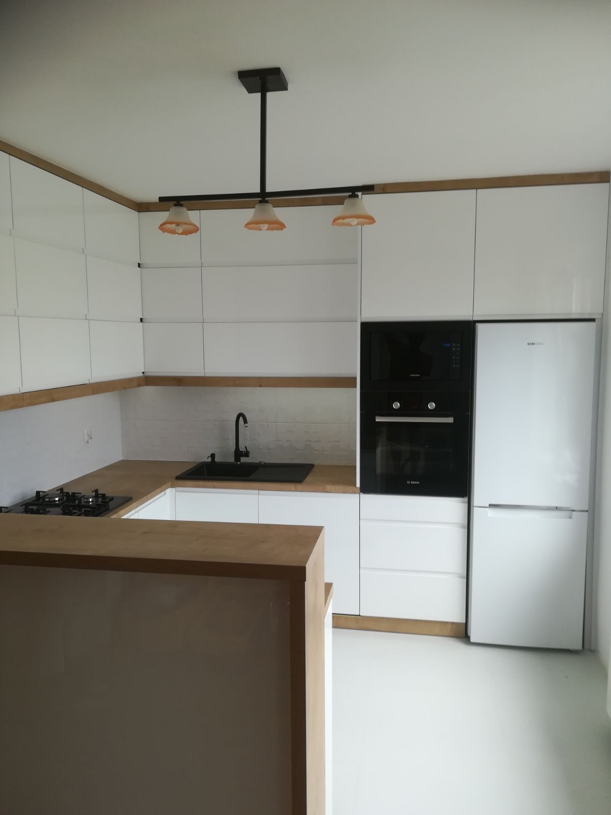 Nowoczesna, biała kuchnia z drewnianymi akcentami, czarnym zlewem i płytą gazową, oświetlona lampą z trzema kloszami w stylu retro.