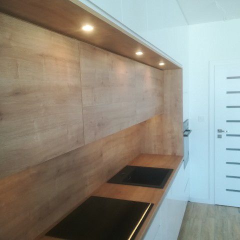 Nowoczesna kuchnia z drewnianymi frontami szafek i czarnym zlewozmywakiem wpuszczanym w blat, oświetlenie punktowe podszafkowe, minimalistyczny design, widoczny fragment płyty indukcyjnej.