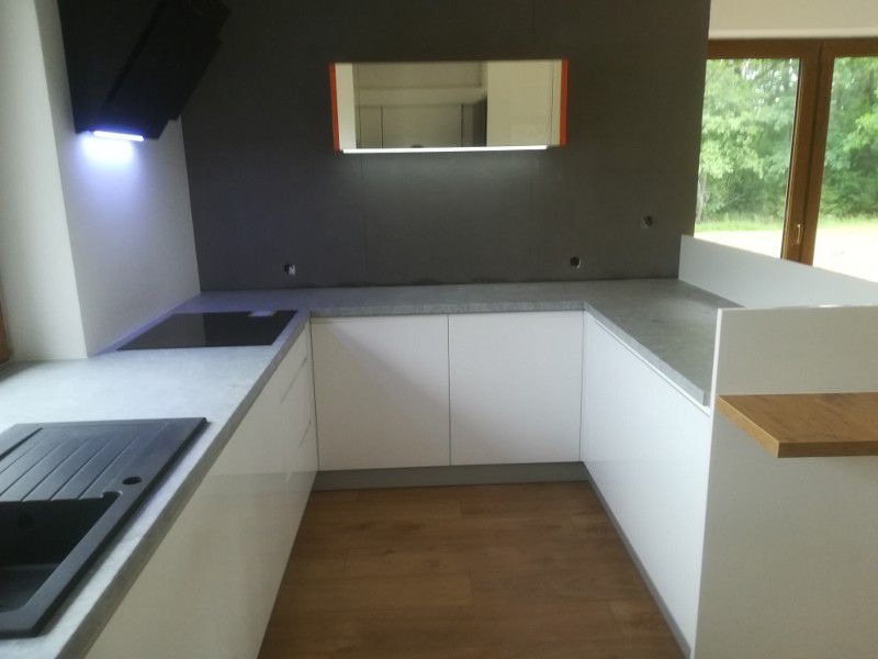 Nowoczesna kuchnia w kształcie litery U z białymi, lakierowanymi frontami, szarym blatem i drewnianą podłogą. Nad płytą grzewczą zamontowany czarny okap z niebieskim podświetleniem LED. Na ścianie...