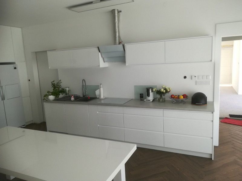 Minimalistyczna, biała kuchnia z frontami bez uchwytów, szarym blatem i podłogą ułożoną w jodełkę. Na blacie misa z owocami i kwiaty.