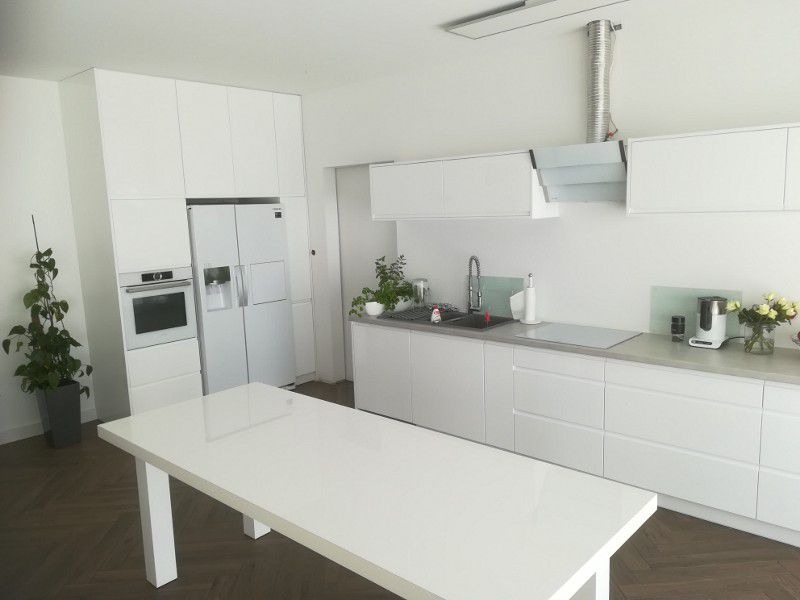 Nowoczesna, minimalistyczna kuchnia w bieli z drewnianą podłogą w jodełkę. Białe, gładkie fronty szafek, zabudowana lodówka i piekarnik. Na blacie zlewozmywak, płyta indukcyjna i drobne akcesoria...