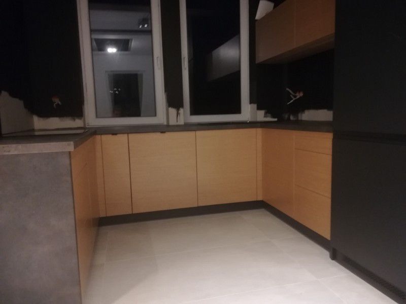 Fragment kuchni w trakcie montażu: widoczne jasne drewniane fronty szafek dolnych, szara ściana z widocznymi śladami po instalacji, białe płytki podłogowe i okna z widokiem na wnętrze innego...