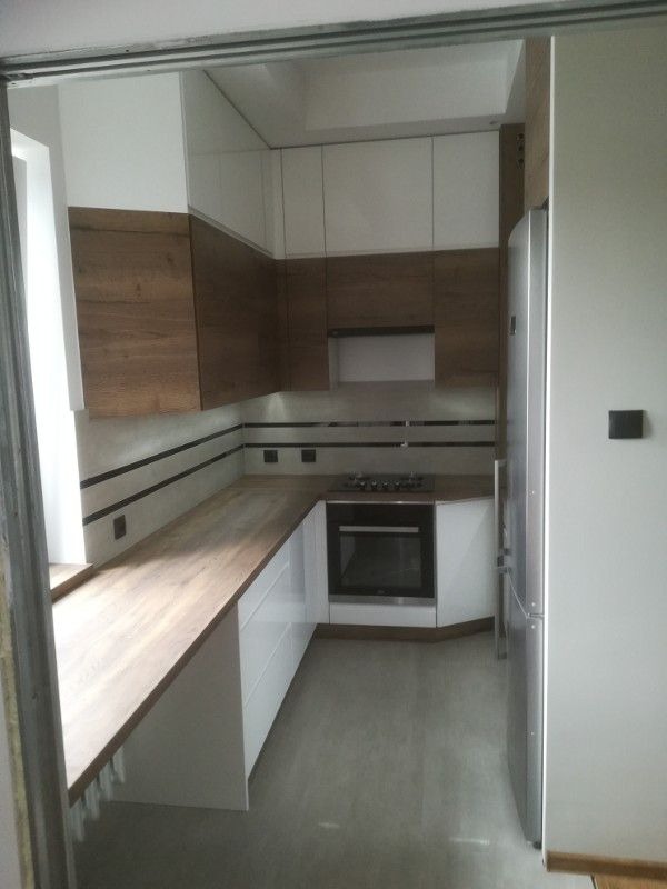 Nowoczesna kuchnia z białymi szafkami górnymi i drewnopodobnymi dolnymi, z blatem w kolorze jasnego drewna, widziana z perspektywy wejścia przez przesuwne drzwi.