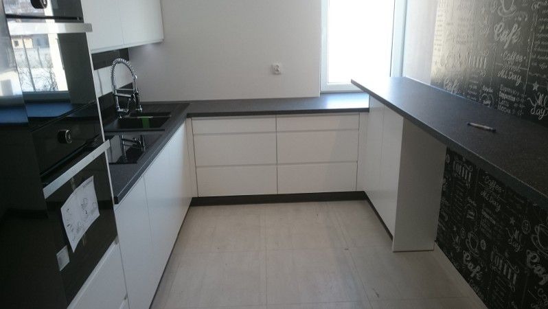 Nowoczesna, minimalistyczna kuchnia z białymi frontami szafek i czarnym blatem, uzupełniona o ścianę z motywem kawowym i niewielki barek.