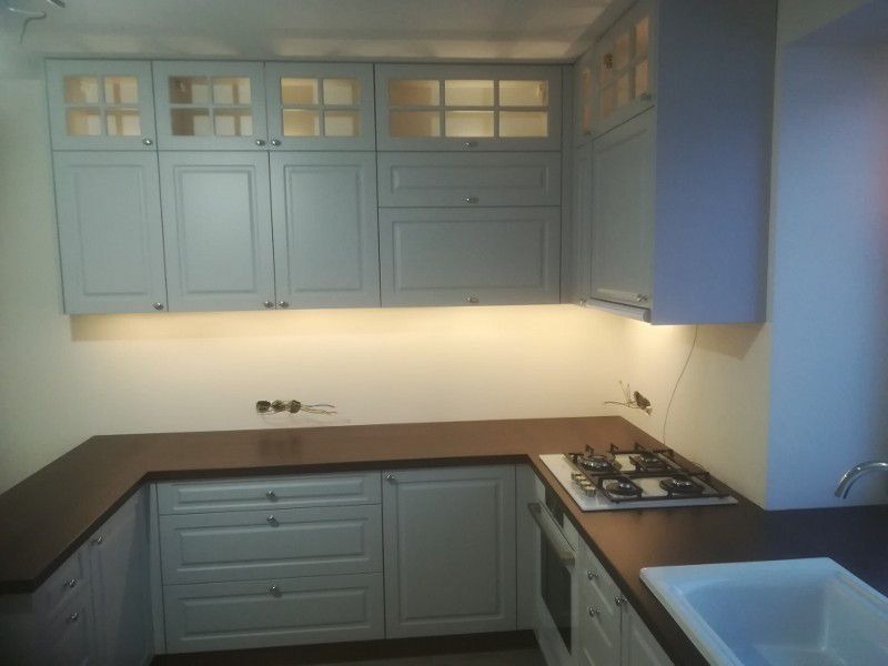 Jasnoszare szafki kuchenne z przeszklonymi górnymi frontami i drewnianym blatem, podświetlane od spodu, z widoczną płytą gazową i niedokończonymi instalacjami elektrycznymi.