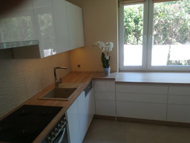 Nowoczesna kuchnia z białymi, lakierowanymi frontami, drewnianym blatem i płytkami z falistym wzorem na ścianie, z oknem i orchideą.