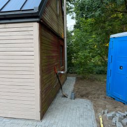 Usługi Brukarskie Wojciech Skwarek - Renowacja drewnianego budynku: świeżo pomalowana beżowa elewacja kontrastuje z brązową, pokrytą mchem. Obok nowo ułożony chodnik z szarej kostki brukowej, narzędzia i niebieska toaleta.