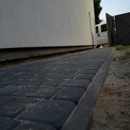 Usługi Brukarskie Wojciech Skwarek - Niskie ujęcie nowo ułożonej ścieżki z ciemnoszarej kostki brukowej z widocznym obrzeżem, biegnącej wzdłuż białej ściany budynku, z piaskiem w fugach.