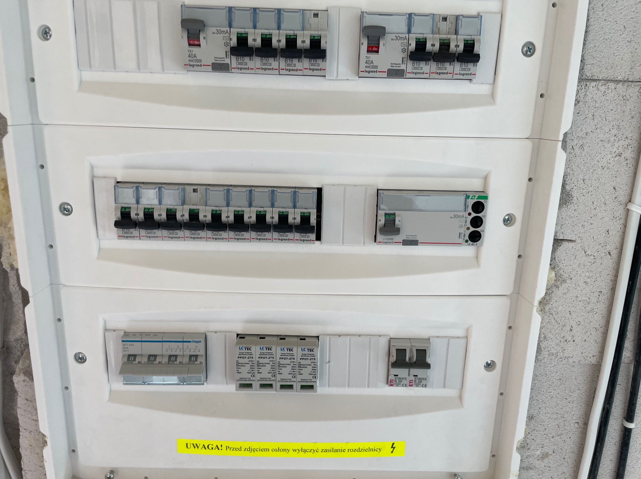 Rozdzielnica elektryczna z zabezpieczeniami różnicowoprądowymi i nadprądowymi, widoczne elementy marki Legrand i Hager, żółta naklejka ostrzegawcza.