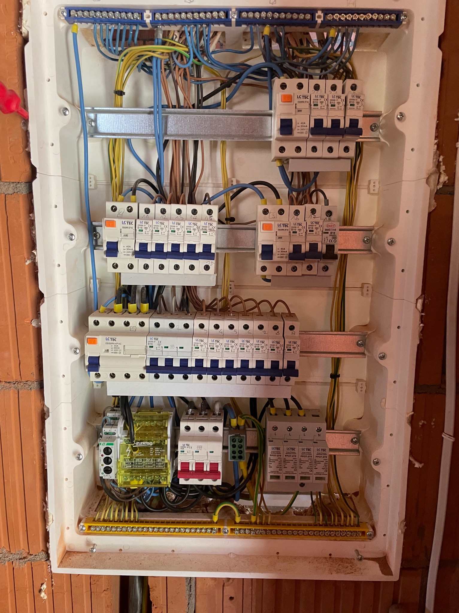 Rozdzielnica elektryczna z zamontowanymi bezpiecznikami i przewodami, widoczna na tle ściany z cegły. Elementy instalacji elektrycznej wewnątrz obudowy.
