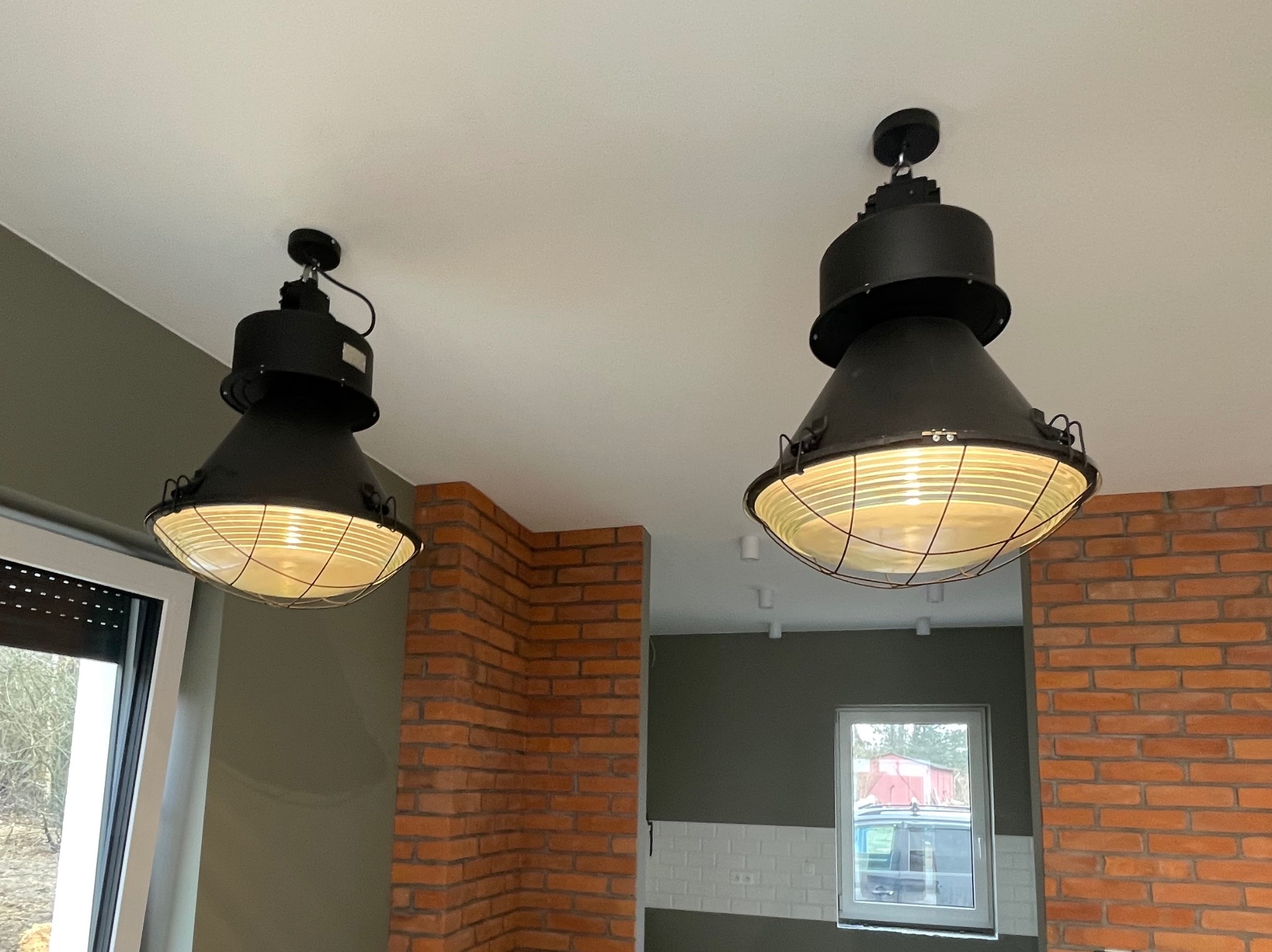 Dwa industrialne, czarne klosze lamp wiszących z żarówkami w ciepłej tonacji, zamontowane na suficie w pomieszczeniu z ceglaną ścianą i oknem.