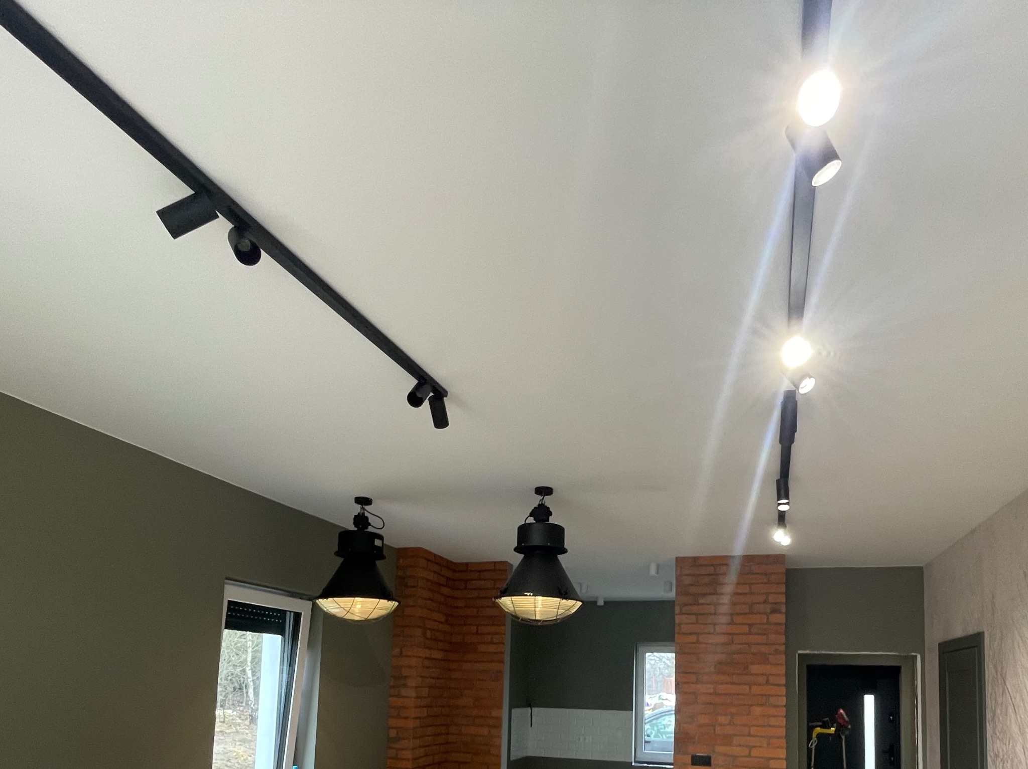 Nowoczesne oświetlenie sufitowe: czarne szyny z reflektorami i industrialne lampy wiszące w pomieszczeniu z ceglaną ścianą i oknem. Jasne światło.