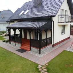 Eurodom Mateusz Hołota - Zadaszony taras przy domu jednorodzinnym z czarną dachówką, widok z góry. Czerwona kostka brukowa tworzy ścieżkę wzdłuż trawnika.