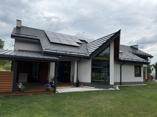 Nowoczesny dom szkieletowy z panelami słonecznymi na dachu i drewnianą pergolą, widok z zewnątrz w pochmurny dzień. Charakterystyczne okno szczytowe.