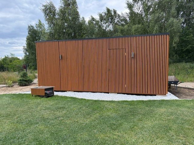 Nowoczesny domek narzędziowy z drewnianą elewacją w pionowe listwy, oświetleniem zewnętrznym i otoczeniem z białego żwiru na tle zielonego trawnika.