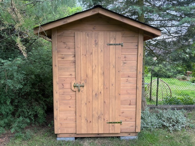 Drewniany domek narzędziowy z czarnym dachem, zamykany na zasuwkę, na betonowych bloczkach, otoczony zielenią. Widok z przodu.