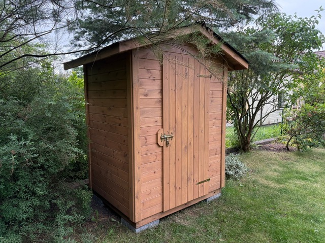 Drewniany domek narzędziowy z zasuwanymi drzwiami, otoczony zielenią ogrodu, z widocznym zadaszeniem i metalowymi okuciami.