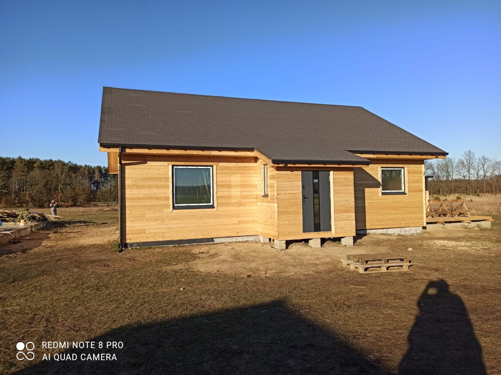 Dom szkieletowy z ciemnym dachem i jasną elewacją drewnianą, widoczny z zewnątrz w słoneczny dzień. Uwagę zwraca minimalistyczny styl i otoczenie w stanie surowym.