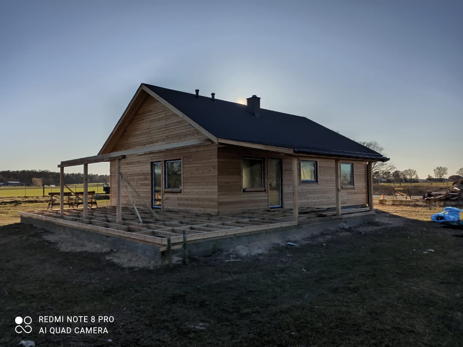 Dom szkieletowy w trakcie budowy, z widoczną konstrukcją drewnianą tarasu. Ściany obłożone deskami, okna wstawione, czarny dach. W tle pola uprawne i drzewa.