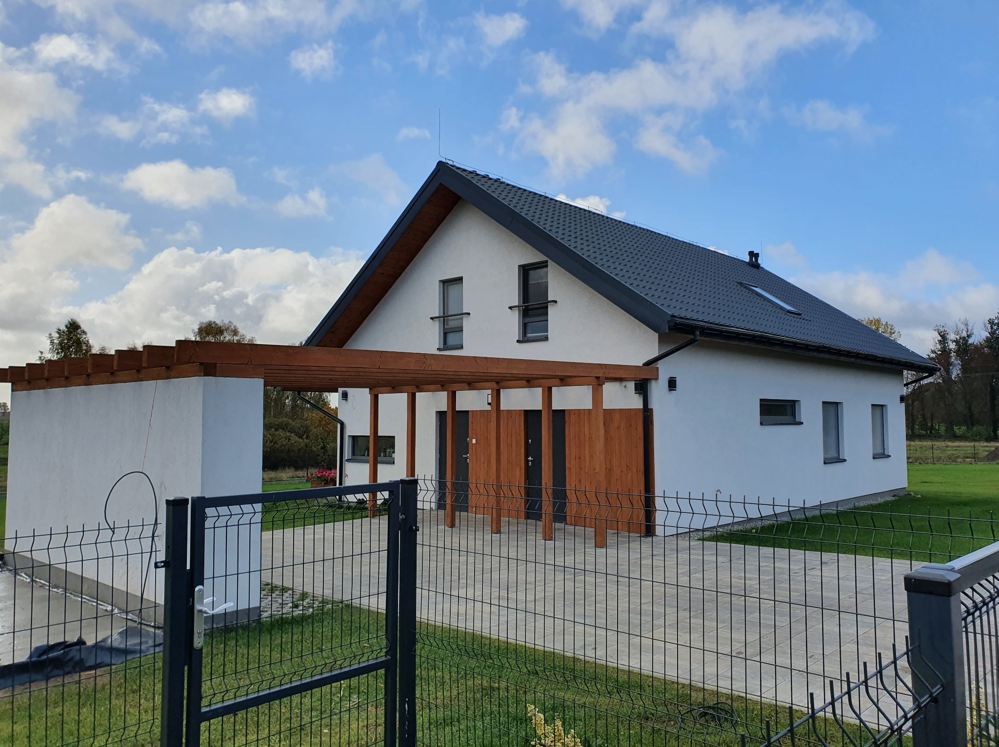 Biały dom szkieletowy z ciemną dachówką i drewnianą pergolą, widok zza ogrodzenia panelowego, błękitne niebo z obłokami w tle.