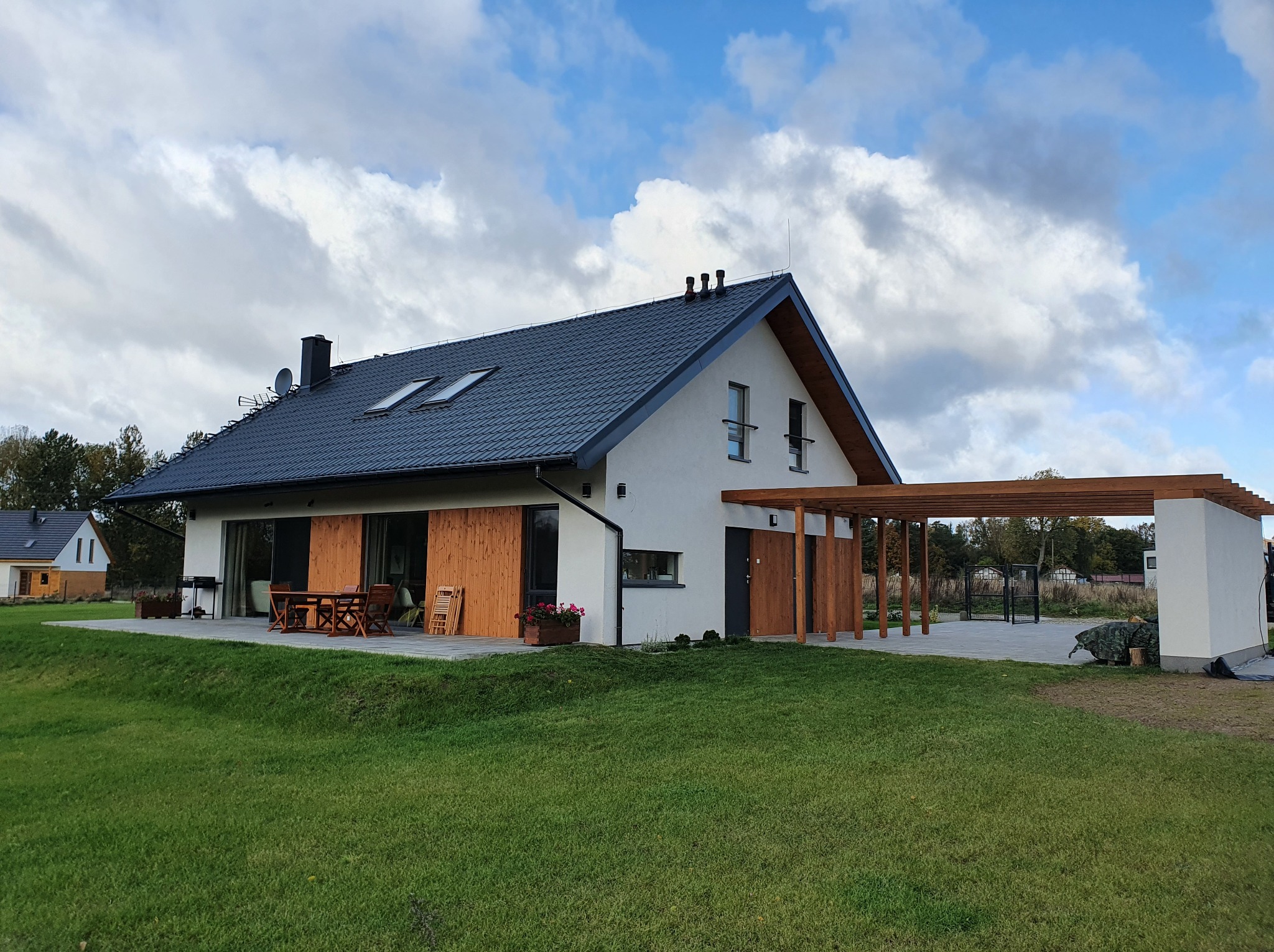 Nowoczesny dom szkieletowy z ciemną dachówką i drewnianą pergolą, widok z ogrodu w pochmurny dzień. Na tarasie drewniane meble ogrodowe.