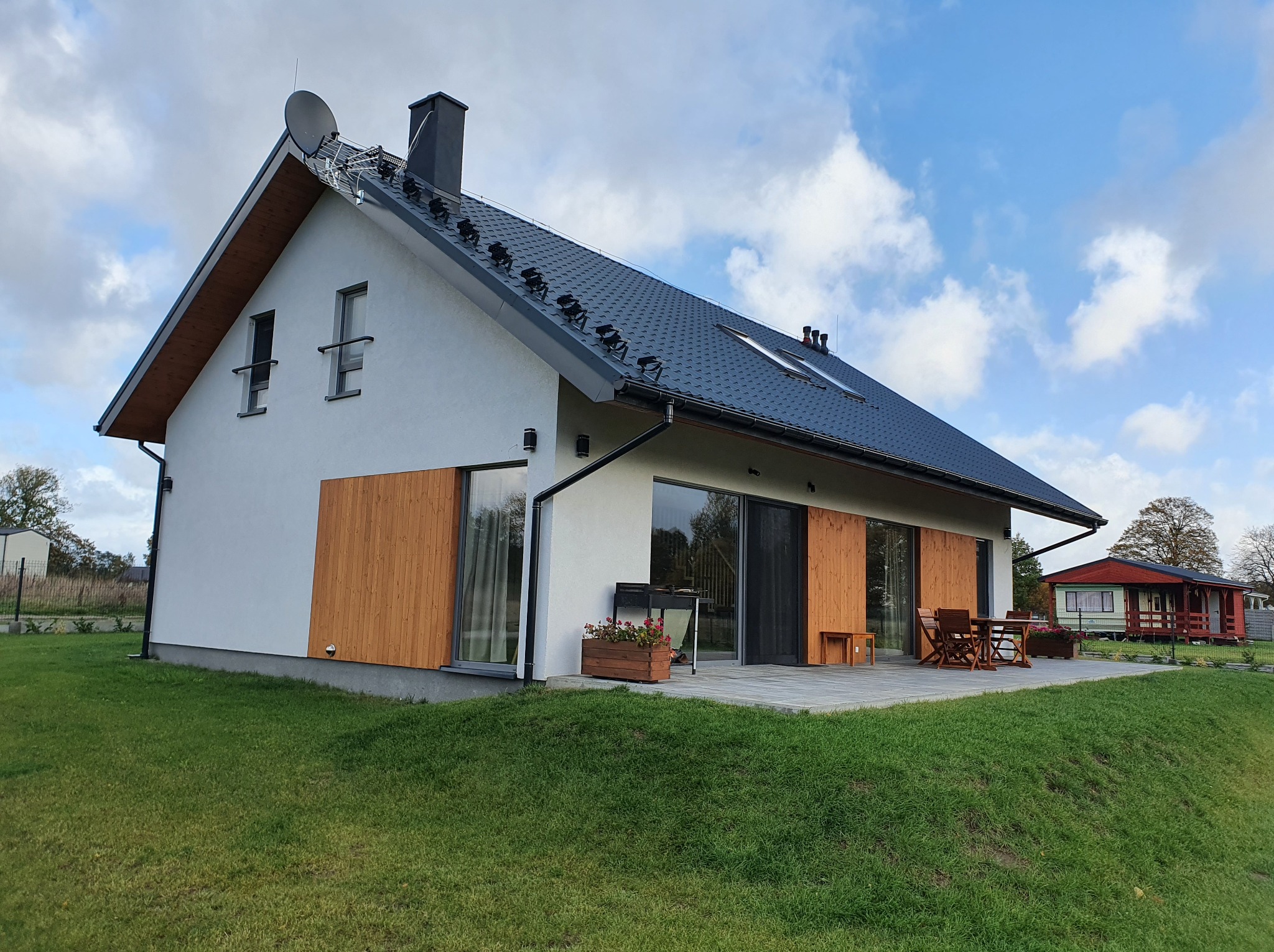 Nowoczesny dom szkieletowy z ciemną dachówką, drewnianymi akcentami i tarasem z meblami ogrodowymi, na tle błękitnego nieba z obłokami.